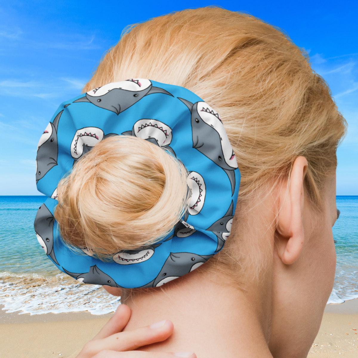 Bright blue scrunchie with smiling gray shark dumplings, worn around a blonde hair bun against a beach background.