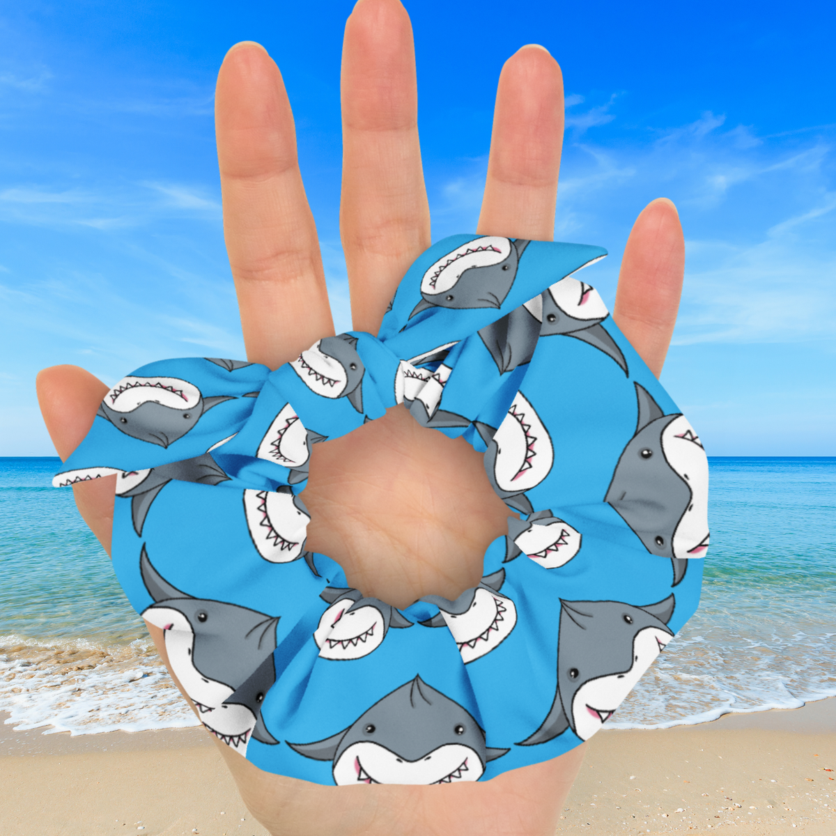 Close-up of a blue shark dumpling bow scrunchie resting on an open hand, showing gathered fabric and smiling gray sharks with white teeth.