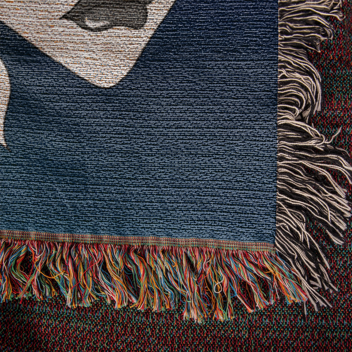 Close-up of 100% cotton woven blanket showing textured yarn construction, dark blue artwork and colorful fringe.