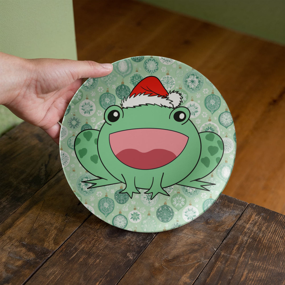 A hand holds a round decorative plate featuring a green cartoon frog wearing a red Santa hat, set against a patterned background with holiday ornaments. The plate is shown on a dark wooden table with a wooden floor and green walls in the background.