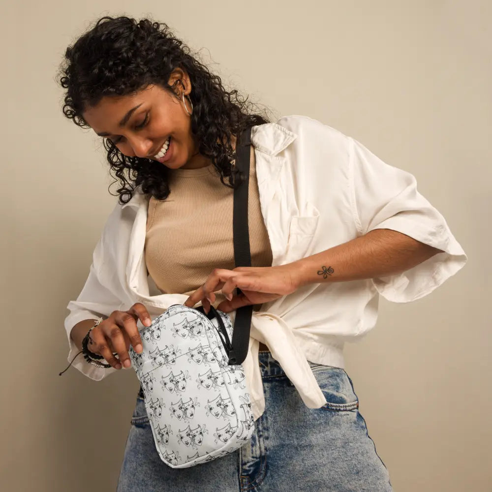 A woman with curly dark hair wearing a casual beige top and white overshirt is smiling as she adjusts the zipper of a white crossbody bag. The bag features a repeating black-and-white theatre mask design symbolizing comedy and tragedy. The woman is styled casually with light blue jeans, and the black adjustable strap of the bag complements her relaxed yet stylish look.