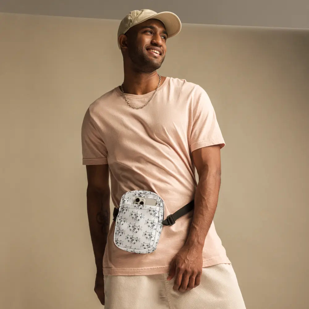 A man wearing a light pink t-shirt, beige pants, and a beige cap is smiling while wearing a white crossbody bag around his waist. The bag features a black-and-white theatre mask design, symbolizing comedy and tragedy. The bag is secured with a black adjustable strap and has a phone visibly placed in the front pocket, showcasing both its practicality and artistic design. The man exudes a casual, modern style.