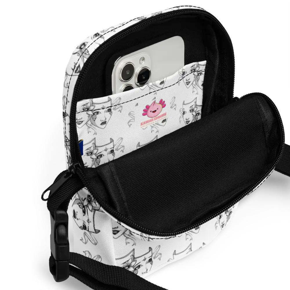 Close-up of a white crossbody bag with a black-and-white theatre mask design, showing the interior pocket holding a smartphone. The inside of the bag features a small branded label with a pink axolotl illustration. The two-way zipper is partially open, revealing the functional storage space, and the black interior contrasts with the artistic exterior design.