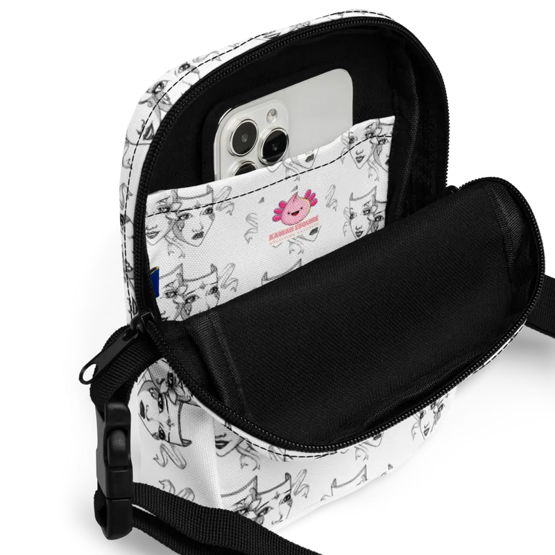 Close-up of a white crossbody bag with a black-and-white theatre mask design, showing the interior pocket holding a smartphone. The inside of the bag features a small branded label with a pink axolotl illustration. The two-way zipper is partially open, revealing the functional storage space, and the black interior contrasts with the artistic exterior design.