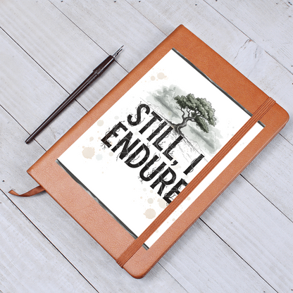 “Still, I Endure” vegan leather journal with tree artwork on the cover, featuring black typography and inspirational roots design, resting on a brown faux leather notebook with elastic band and ribbon bookmark.
