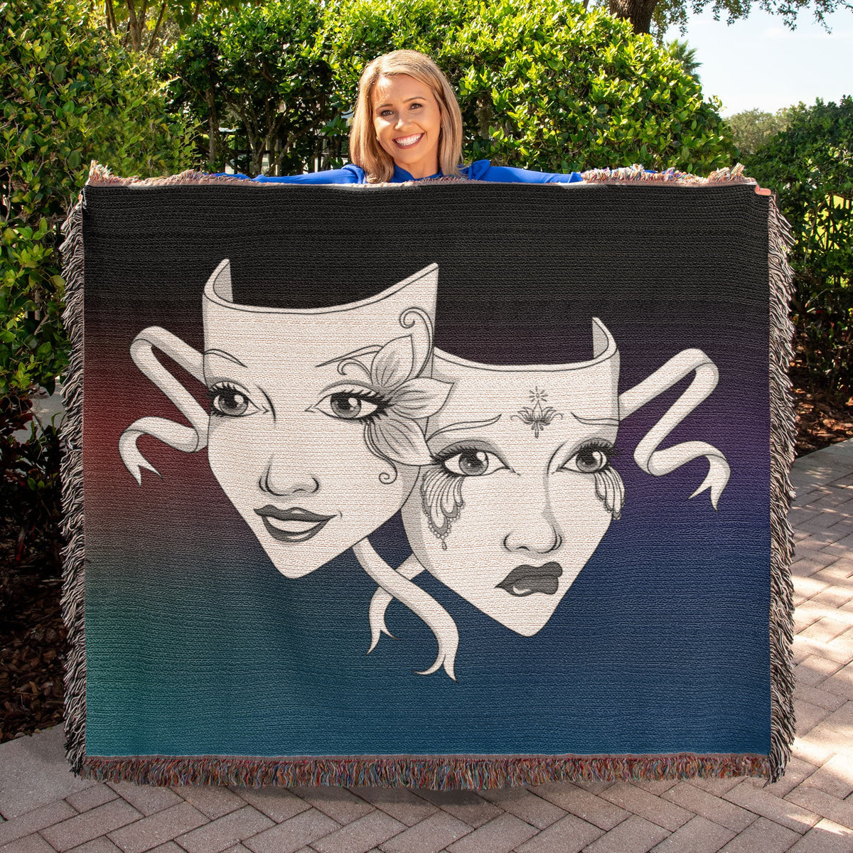 Woman holding large theatre masks woven blanket featuring comedy and tragedy artwork with colorful cotton yarn fringe.